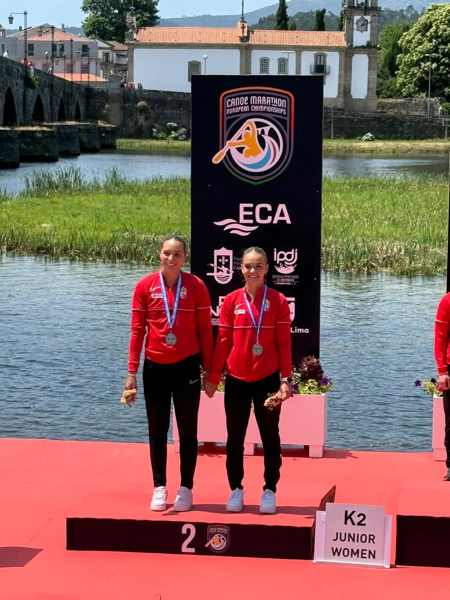 Két magyar női kajakos piros melegítőben áll a dobogó második fokán, nyakukban ezüstérem, a K2 junior női kategória eredményhirdetésén a maraton EB-n.