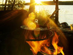 Pfanne mit frisch gebratenem Fisch über dem offenen Feuer bei Sonnenuntergang – Outdoor-Küche auf Kajaktour in Finnlands Saimaa-Seengebiet