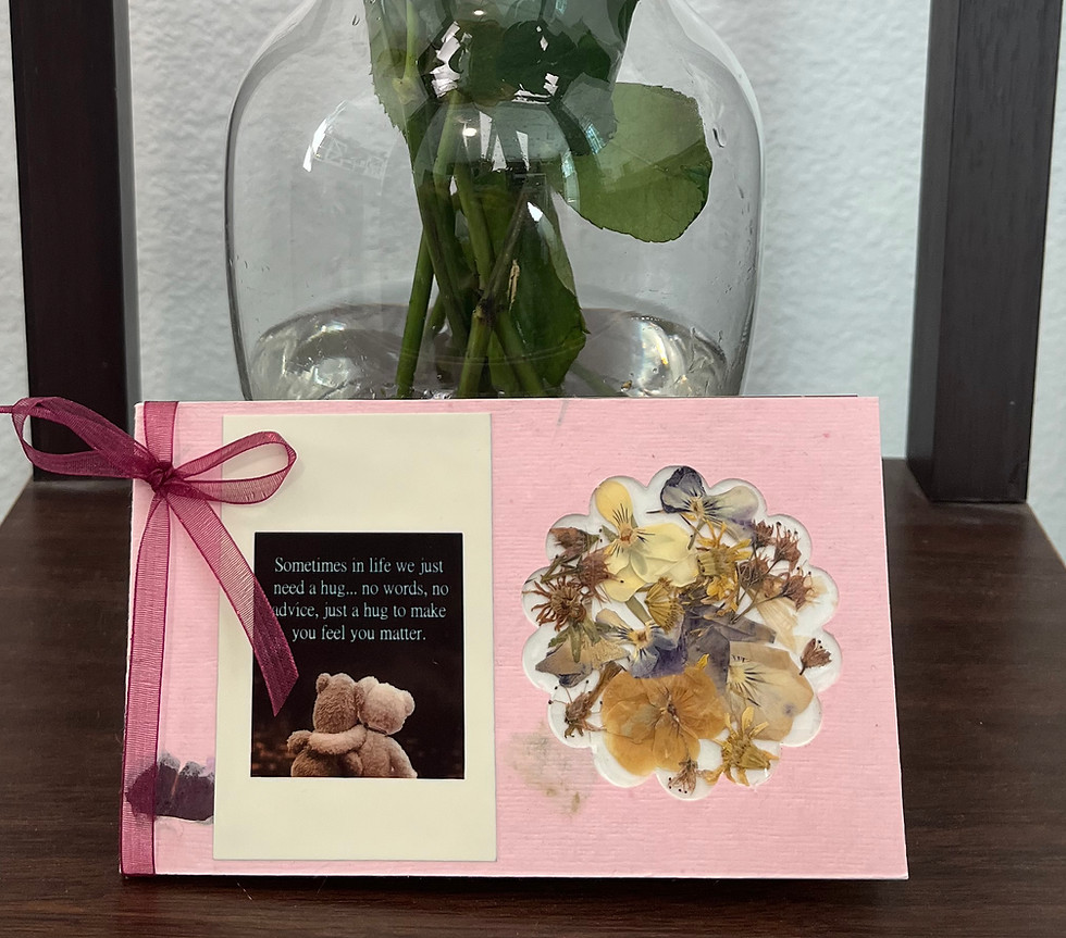 Small pink greeting card. On the left there is a dark magenta ribbon with a bow. On the right side, there is a cut out shape of a flower. Inside the flower cut out is dried flowers. On the left side, there is a photo of the back of two brown teddy bears hugging. Text is on top: "Sometimes in life we just need a hug...no words, no advice, just a hug to make you feel better.