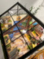 Top down view of a miniature greenhouse that has clear plastic windows around to see the inside. Inside there is a functional ceiling light. There are shelves with plants, boxes with utensils such as a spake, rake, etc. There is also an easel with a painting. There are also paintings and small decorative items. 