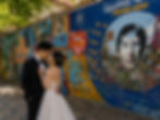 A couple in wedding attire embraces by the colorful Lennon Wall, featuring vivid graffiti and text like "Lennon Wall of Freedom & Energy."
