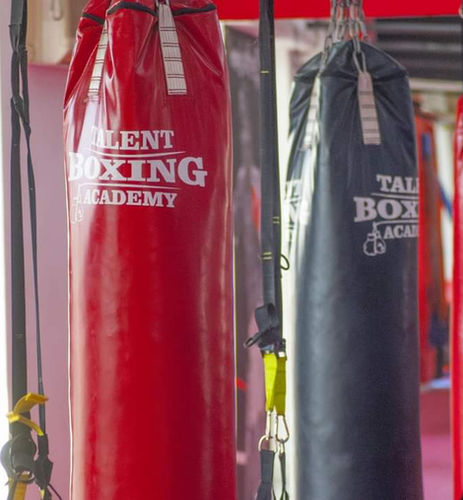 4 punching bags in a row, 3 red and 1 black. They all have the logo of the boxing gym on them that says "Talent Boxing Academy"
