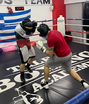 Two men wearing boxing gloves and headgear, boxing inside a ring. There is a Greek flag on the wall and a clock on top
