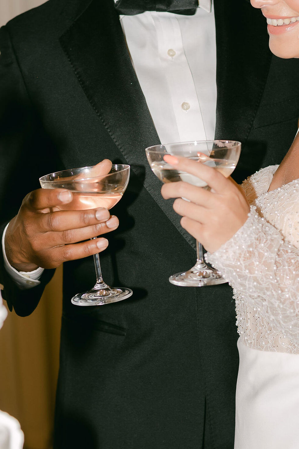 Man and woman in formal attire hold champagne glasses, smiling. Black suit and white lace dress. Elegant and celebratory mood.