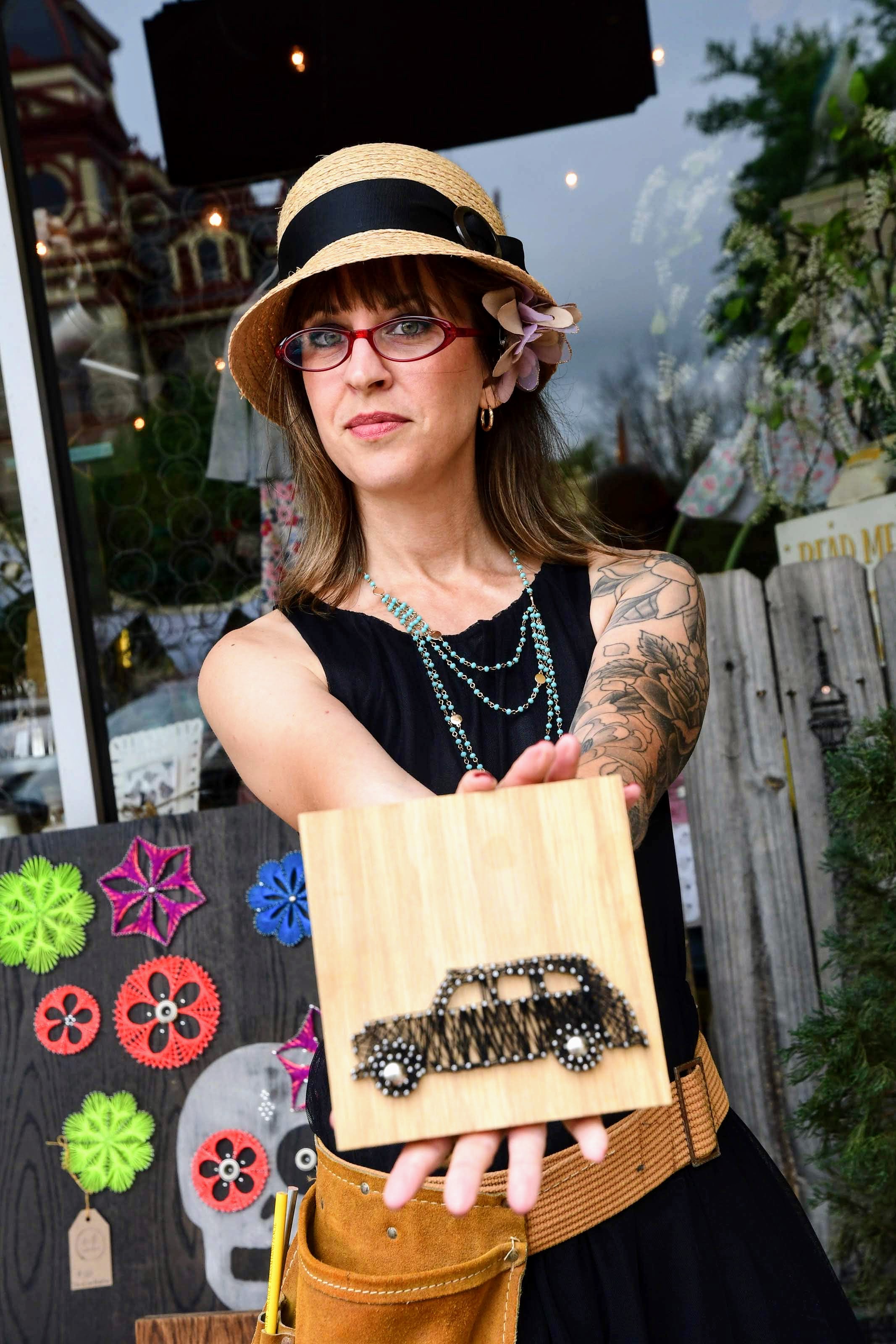 Brandy Schuenemann in a hat outside holding a work of art of a car on wood made of string and nails