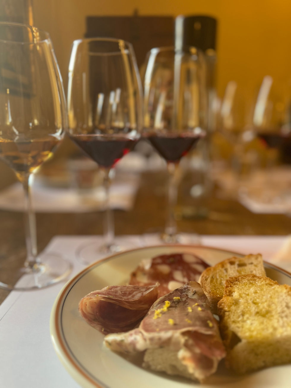 Plate with sliced meats and bread in the foreground, several glasses of red orange and white wine in the background on a wooden table. Warm lighting.