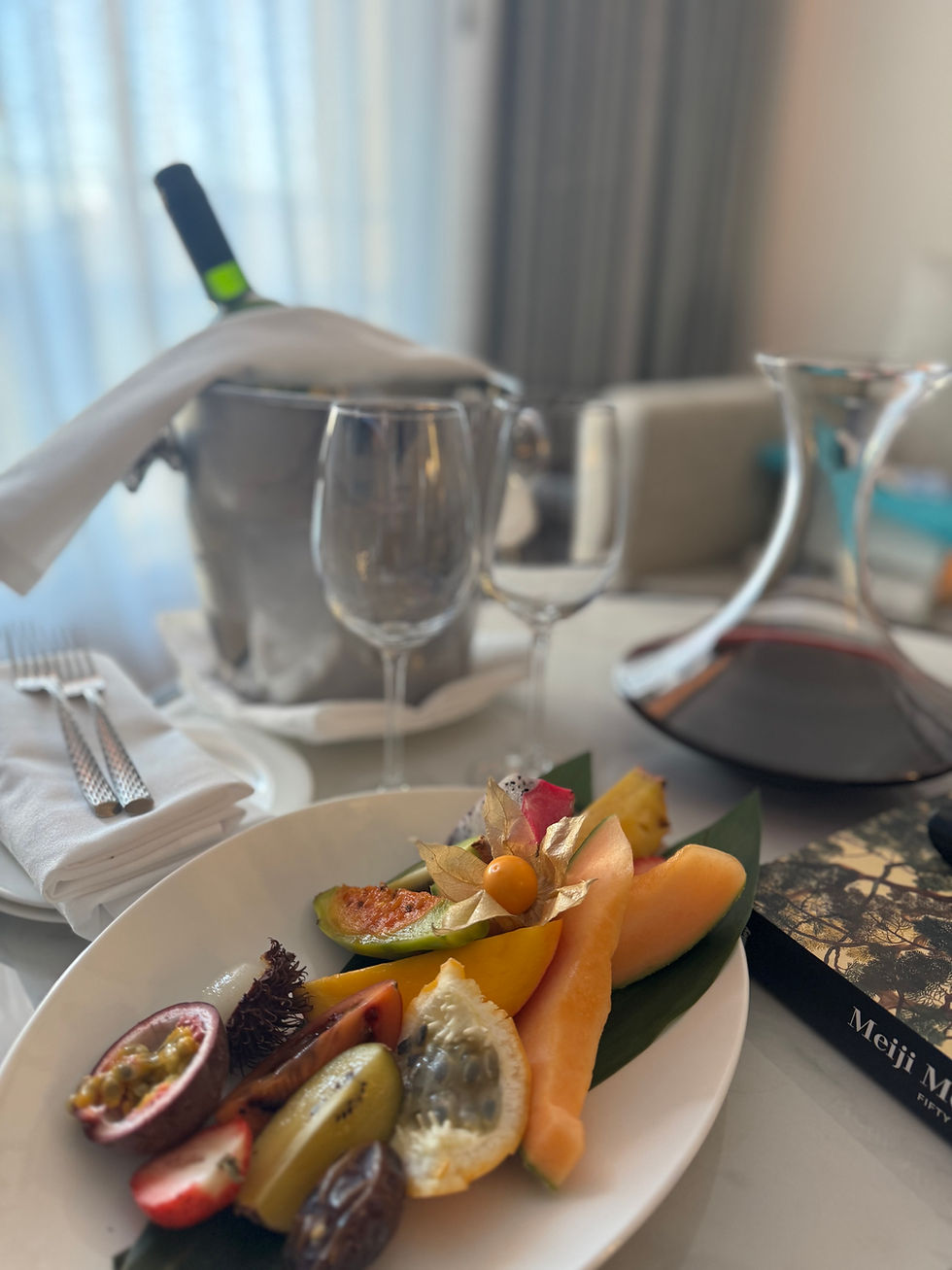 Plate with assorted fruits on a table, a wine bottle in ice, two glasses, decanter, and a book titled "Meiji." Sunlight filters through curtains.