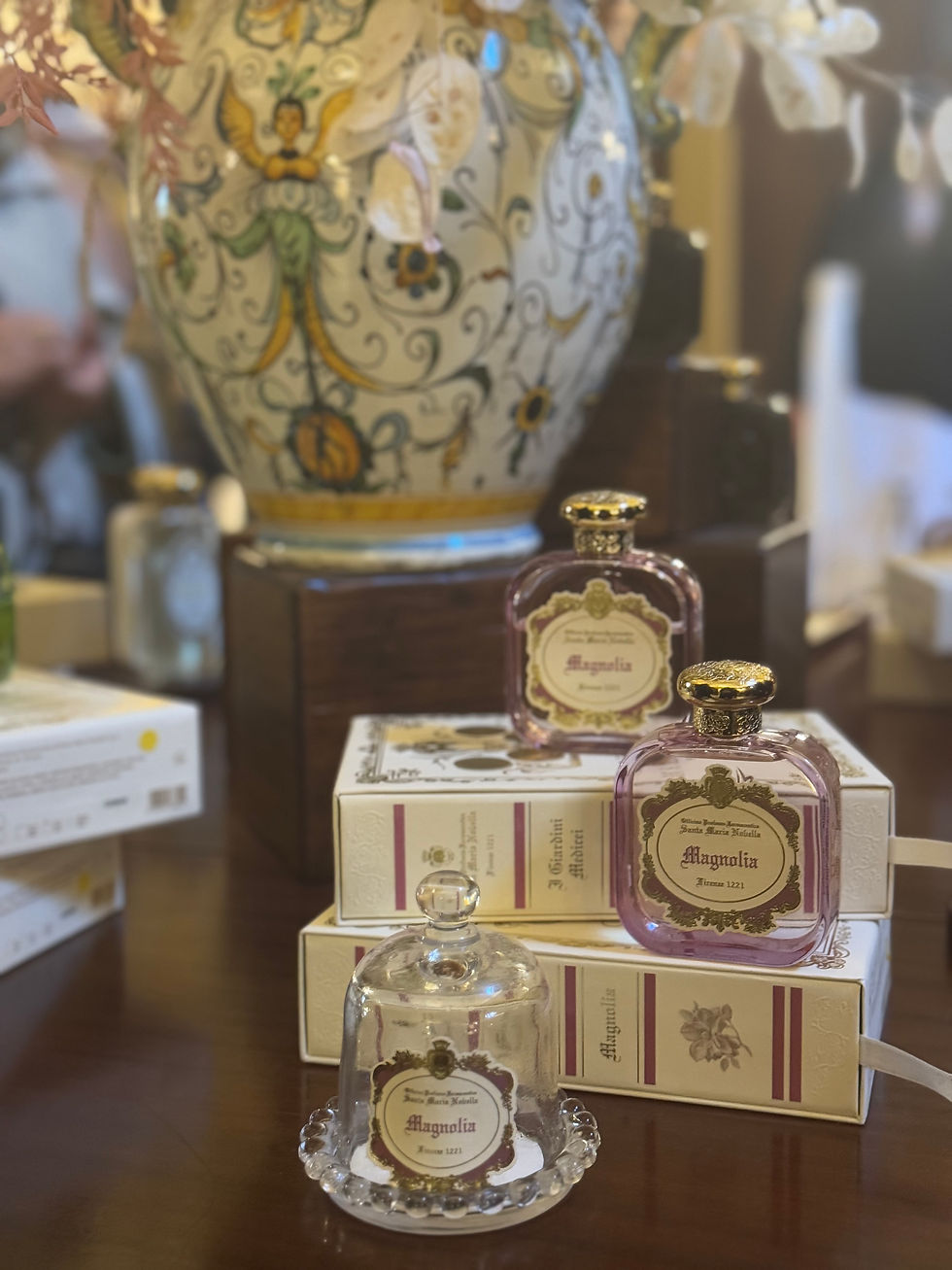 Perfume bottles labeled "Magnolia" on stacked boxes, next to a decorative vase with floral design, set on a wooden table.