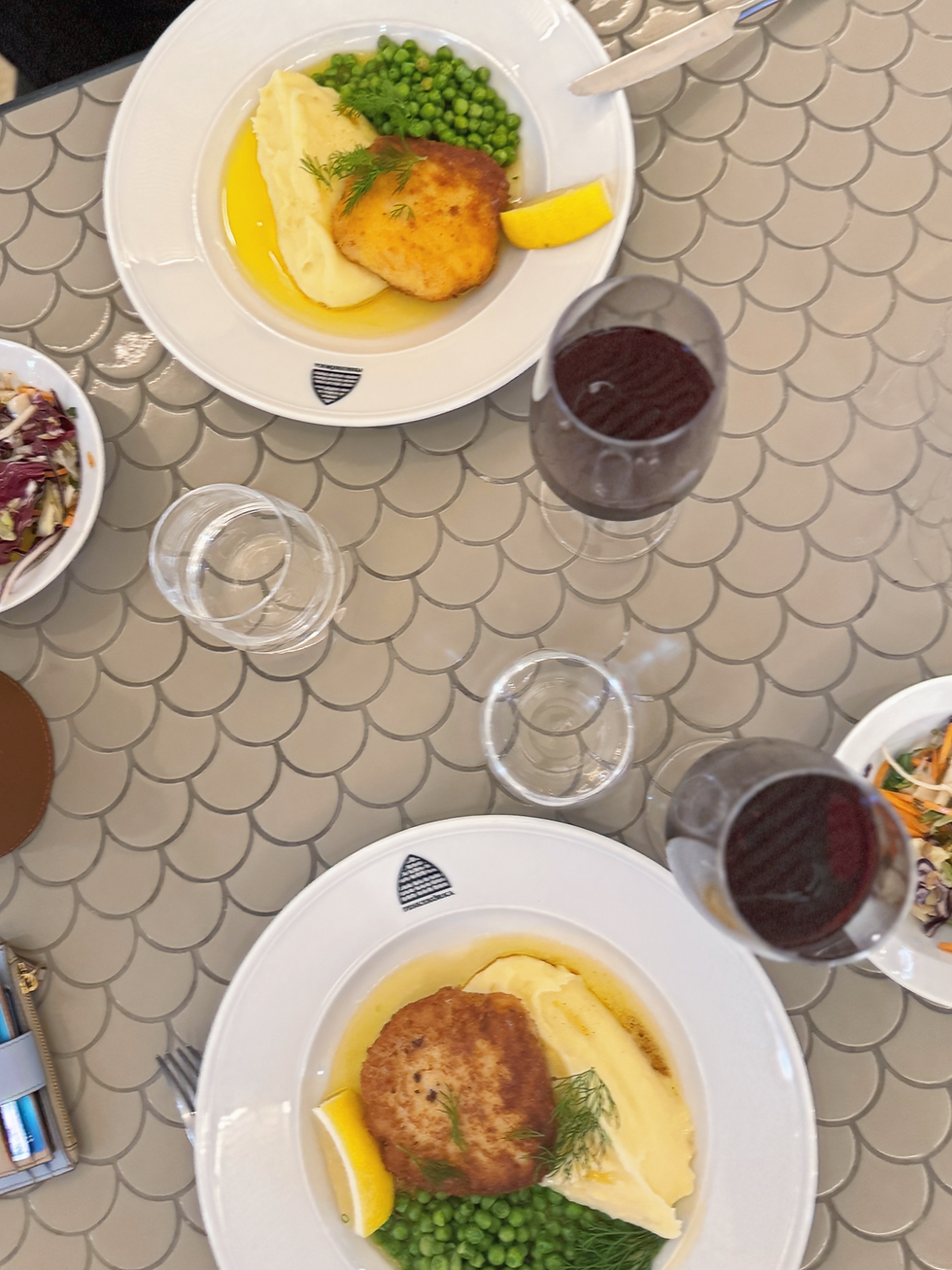 Plates of breaded fishcakes, mashed potatoes, peas, and lemon on a fish scale patterned table with red wine and salad bowls. Relaxed mood.