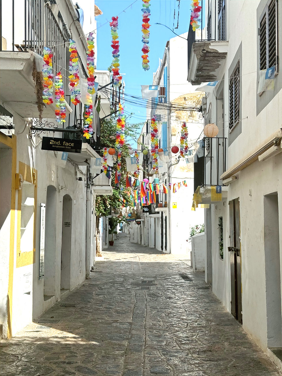 Narrow street with white buildings, colorful flags, and hanging decorations. Sunny day with blue sky. Sign reads "2nd. face."