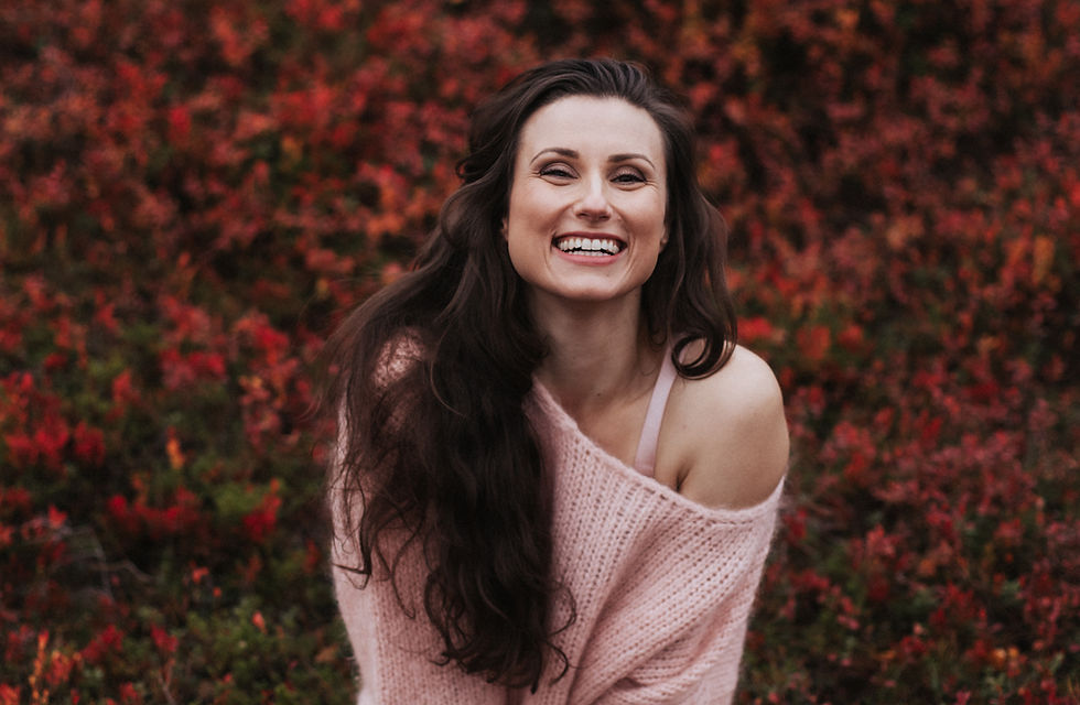 Woman with brown hair sitting in the ground in her boudoir photo smiling and wearing a cardigan in Finnish Lapland. Photo is covered with autumnal colors in Ylläs, Äkäslompolo, Finland.