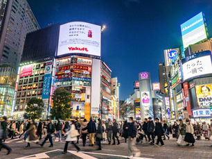 A reflective travel scene in Tokyo showing a busy urban street near Shibuya Crossing, with crowds of people walking in different directions under glowing city lights. The image captures the feeling of being slightly lost yet safe and curious, surrounded by towering buildings, train station signs, and small side streets waiting to be explored.
