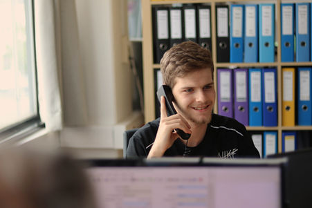 ein junger Mann lächelt und telefoniert mit einem Telefonhörer; hinter ihm bunte Ordner in einem Regal