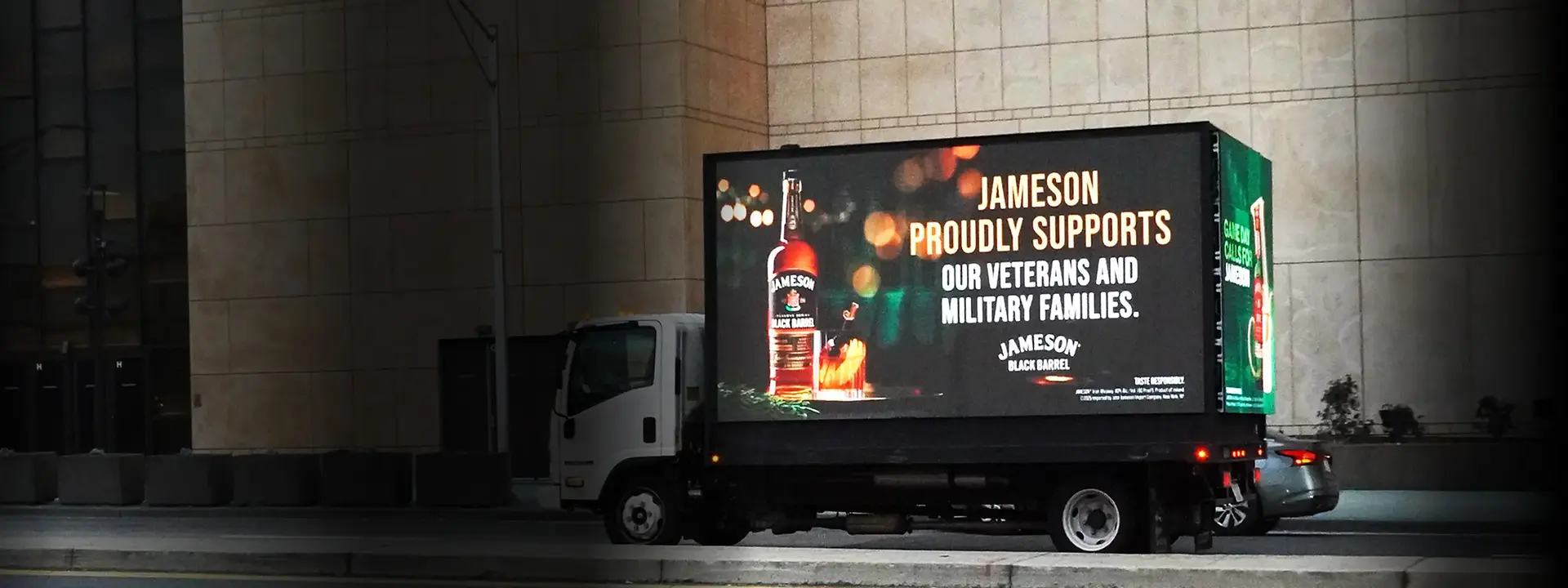 A Jameson Black Barrel campaign runs on an MATV Mobile Billboard parked against a building wall after dark, its dedicated screens carrying the brand's message that Jameson proudly supports veterans and military families. MATV used Stationary Advertising and Transit Advertising to put the beverage brand in front of audiences around sporting events, building Brand Awareness where fans gather.