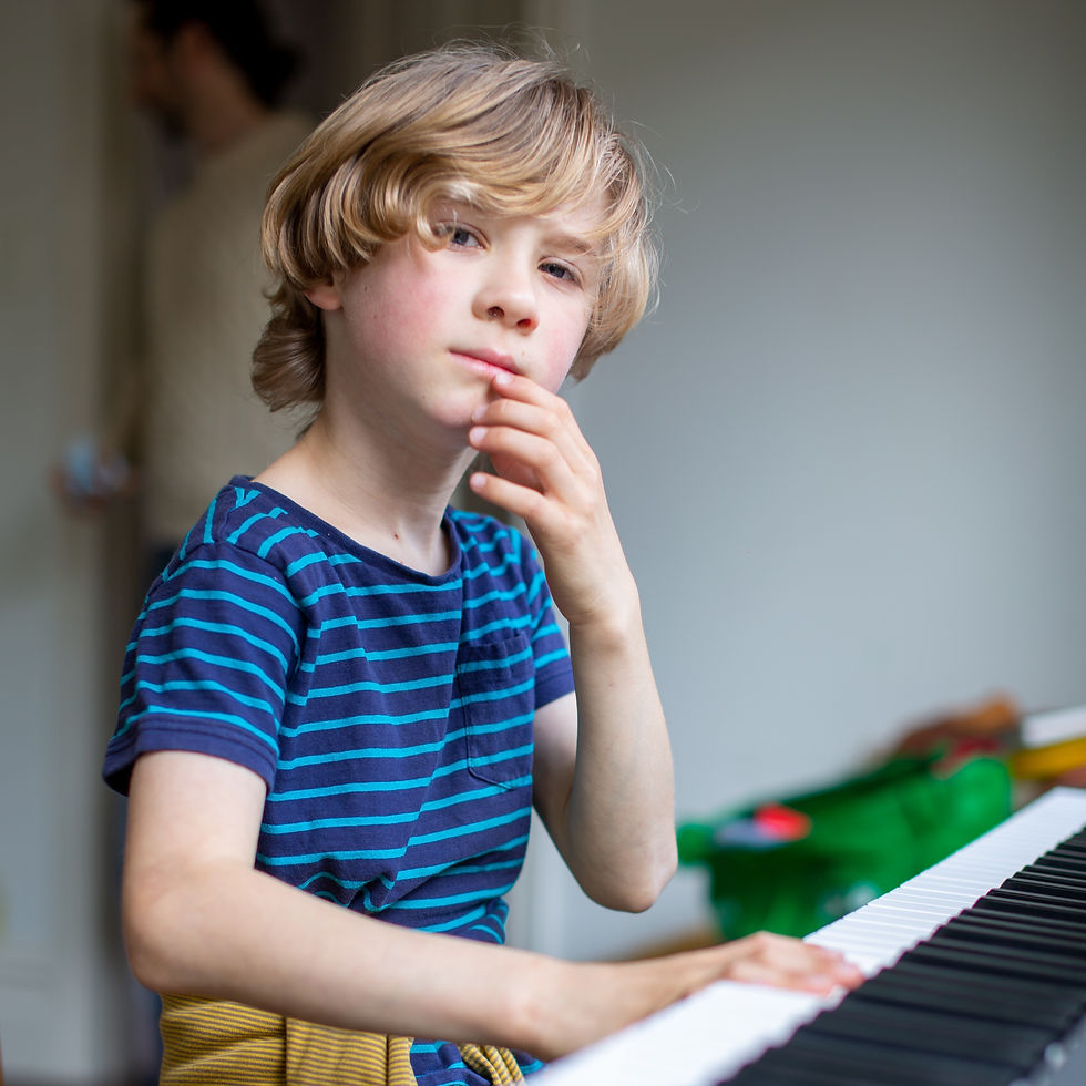 Boy in a striped shirt sits pensively at a keyboard, hand on chin. Blurred figure in the background. Bright, casual indoor setting.
