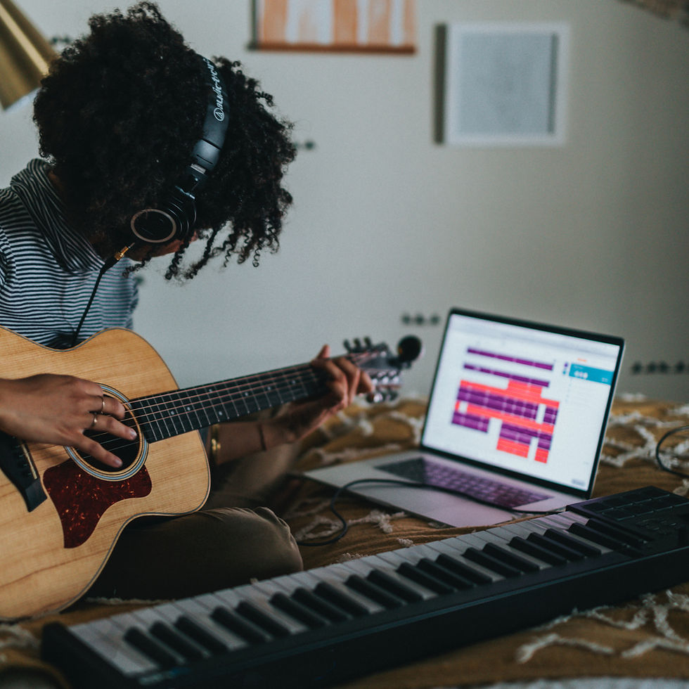 Person in headphones plays acoustic guitar on a bed beside a laptop showing music tracks and a keyboard, focused mood