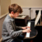 Boy playing piano intently in a cozy room. Sheet music on stand, keyboard in background. He's wearing a gray shirt, focused expression.