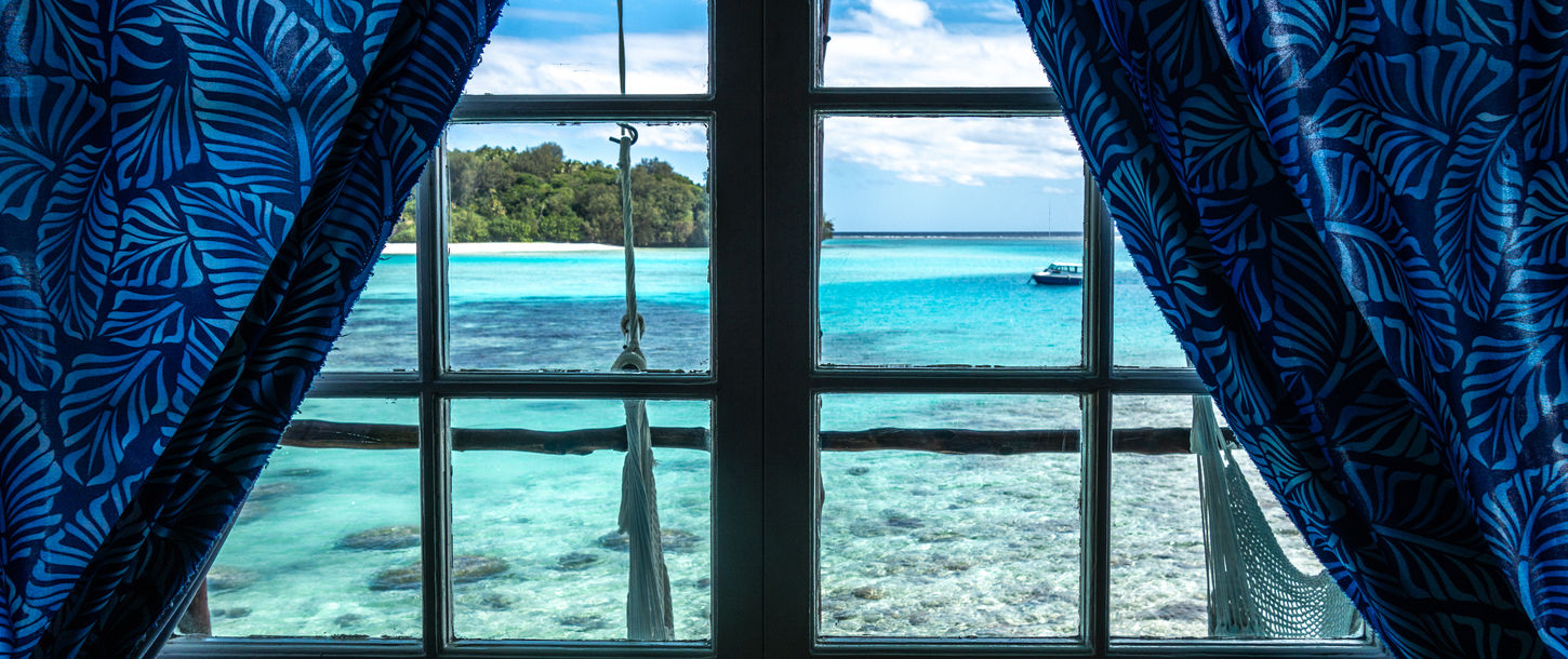 Window view to turquoise ocean, tropical island, boat, blue curtains.