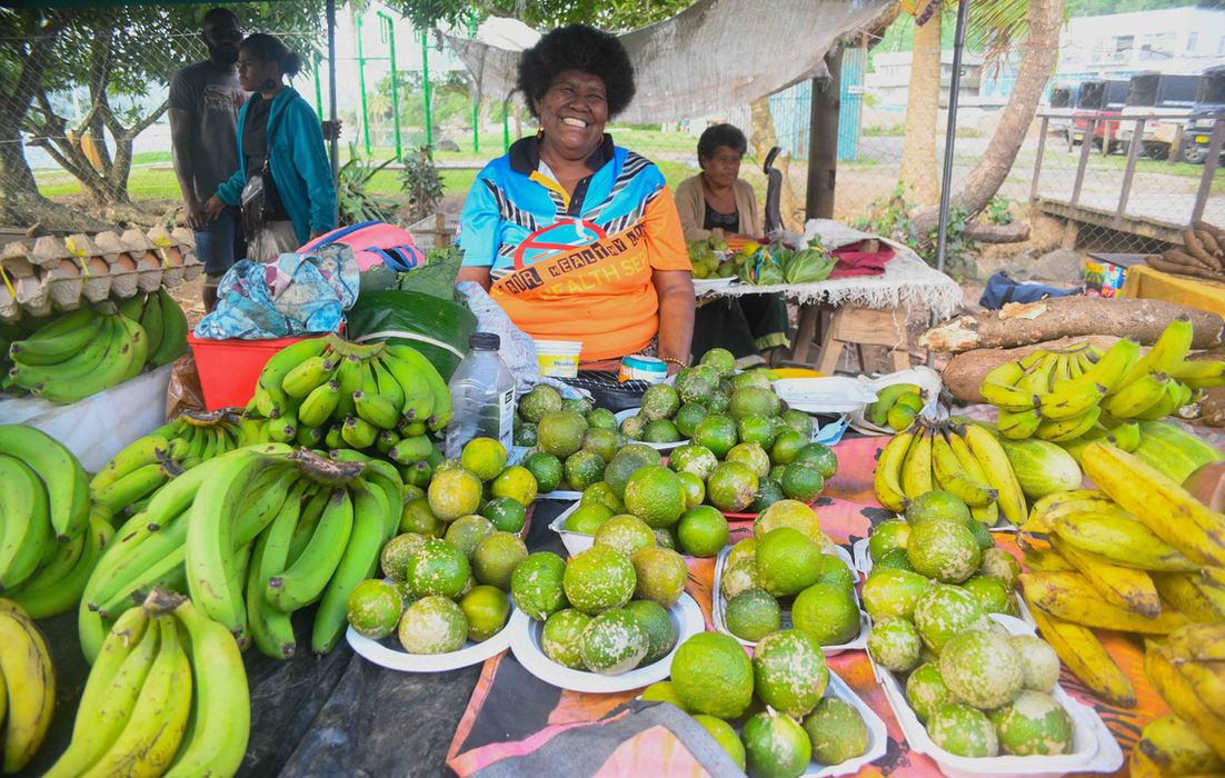 Woman selling fruits smiling at market stall with bananas and limes, Gecko Digital Fiji