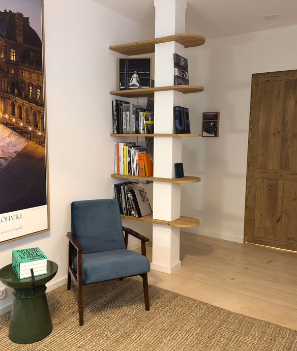 étagères en bois aménagées autour d'un poteau porteur avec au premier plan un fauteuil bleu vintage et un tabouret en céramique de couleur verte