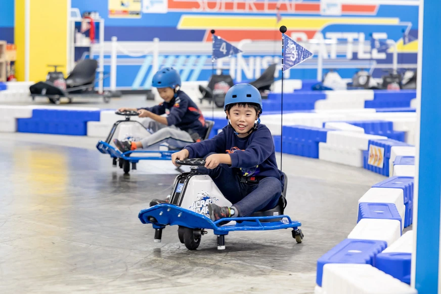 兩個戴藍色安全帽的男孩在室內賽道開卡丁車,表情愉悅。背景為藍白色調,牆上有「FINISH」字樣