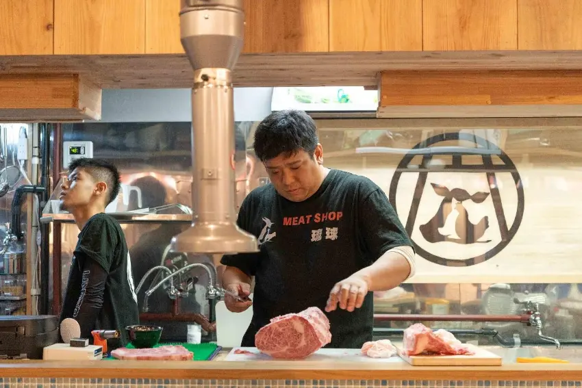 肉舖內兩名男子,一人低頭切割案板上的大塊生肉,另一人站在旁邊;櫃檯前擺著多塊肉,背景有“MEAT SHOP”字樣與店鋪標誌,氛圍專注忙碌