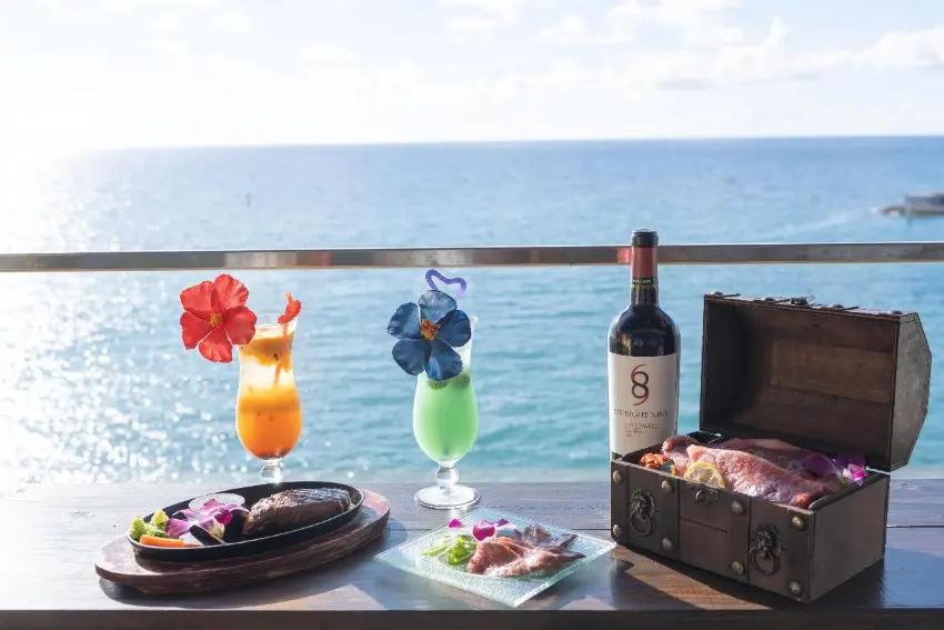 海邊露台上擺著兩杯花飾雞尾酒、一瓶紅酒、牛排餐盤和裝滿食物的木箱,陽光明亮,海景寧靜愜意,瓶身有數字6