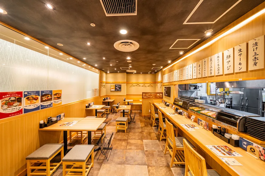 日式餐廳室內,木質桌椅整齊排列,牆上有菜單,地板為暖色瓷磚。內設開放式壽司櫃檯,氣氛溫馨