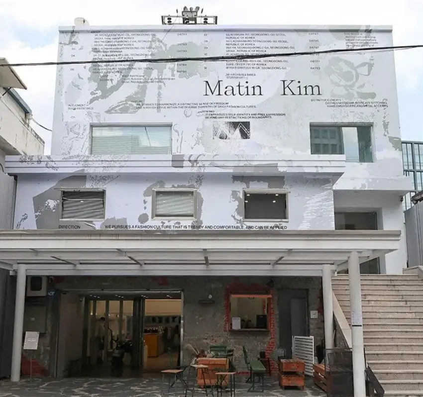 白色外牆的兩層建築,立面寫著 Martin Kim,牆面有多行英文說明;下方是半開放入口、桌椅和樓梯,整體安靜簡潔