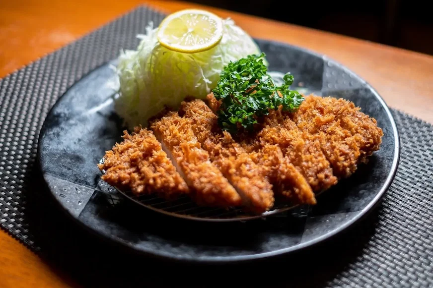 炸豬排配高麗菜和檸檬片,擺放在黑色盤子裡,背景是深色桌布,食物呈現出誘人的金黃色