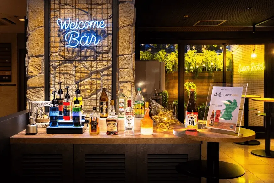 歡迎酒吧,木桌上擺滿各類酒瓶。背景石牆上有「Welcome Bar」霓虹燈,窗外綠植增添生機,氛圍溫馨
