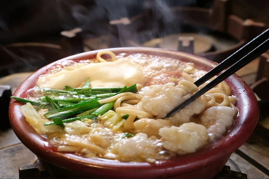 紅色碗中盛滿熱氣騰騰的麵條、青蔥和小塊魚肉。黑色筷子正夾住魚肉,背景為模糊的廚房環境。氣氛溫暖