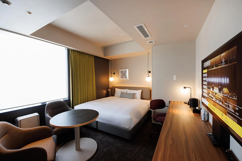 現代飯店房間,內有一張雙人床、靠窗的沙發和圓桌。牆上掛有裝飾畫,大電視螢幕展示飯店資訊,氣氛溫馨