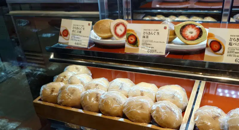 商店櫥窗內展示多種和果子,前排包裝的麵包,後排有草莓夾心的白餅。商品標籤寫有日文,背景為深色