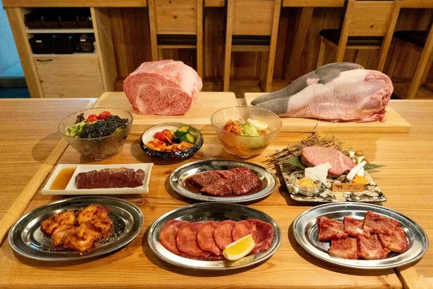 木桌上擺滿和牛拼盤與配菜,後方展示整塊生牛肉和魚肉;前景多盤烤肉、沙拉、醬菜,日式餐廳氛圍豐盛誘人