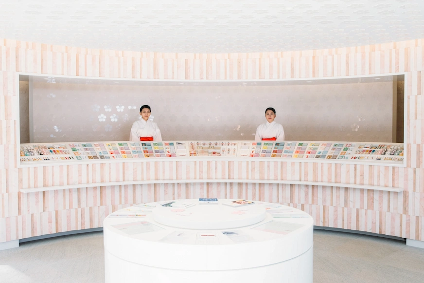 現代化店內,兩位身穿白色和服的女性站在展示櫃後,櫃檯上陳列著各種商品,背景為淡粉色木紋牆,氛圍寧靜