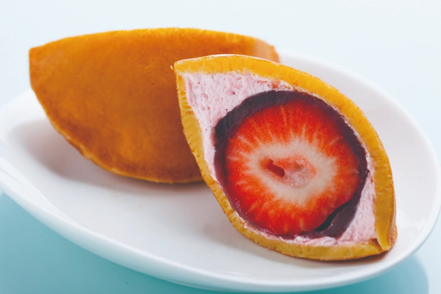 白色盤子裡呈現一塊金黃色的日式甜點,切開展示草莓夾心,粉紅色奶油圍繞,背景為淺藍色,顯得清新誘人