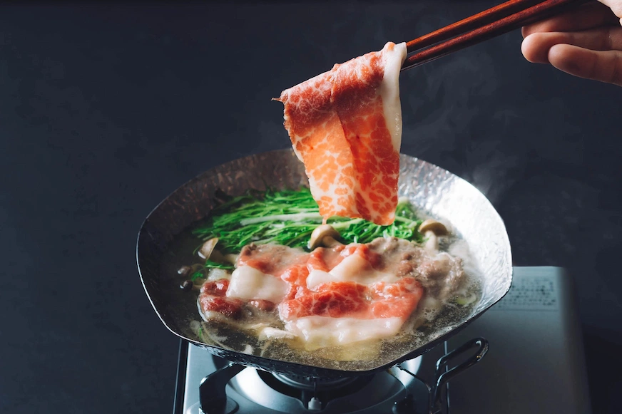 鍋中烹調生鮮食材,背景深色,火鍋熱氣騰騰。有人用筷子夾起一片紅白相間的肉片,旁邊有蔬菜和蘑菇