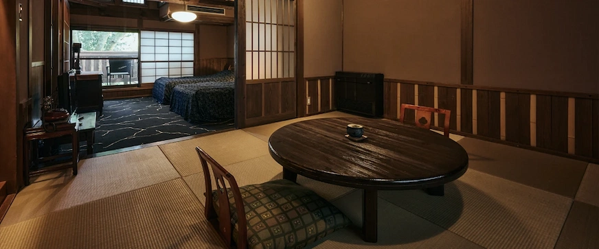 傳統日式房間,中央放置圓形木桌,桌上有茶杯。背景為鋪有藍色床單的床,窗外綠意盎然,整體氛圍寧靜