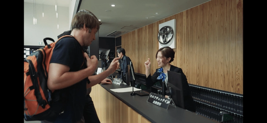 男士背著橘色背包與櫃檯後方的女接待員交流,櫃檯上有標示。背景是木質裝飾。兩人面帶微笑,氛圍輕鬆