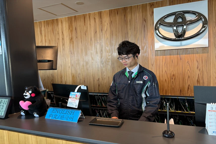 櫃檯上有穿制服的工作人員,背景是木製牆面,牆上有豐田標誌。前景有熊玩偶和語言指示牌,氛圍正式