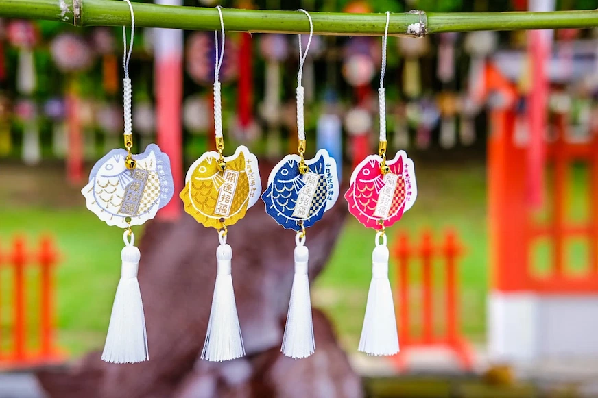 四個不同顏色的魚形掛飾懸掛在竹竿上,分別為白、黃、藍、粉紅色,上有日文,背景為模糊的綠地和紅色建築