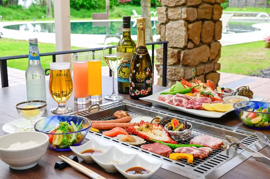 戶外燒烤餐桌上有多種肉類、海鮮、蔬菜,旁有飲品、酒瓶及玻璃杯。背景為綠地與泳池,氛圍輕鬆愉快