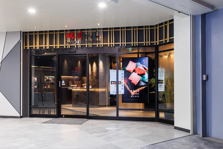 餐廳外觀,入口玻璃門上方有紅色招牌。門旁電子螢幕顯示肉類影像,室內燈光柔和,牆面為深色調設計
