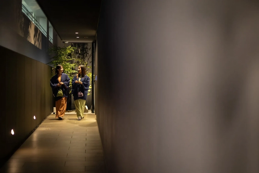 走廊內,兩位穿著深色上衣的女性正微笑交談,背景有綠色植物和暖色燈光,營造出溫馨氛圍