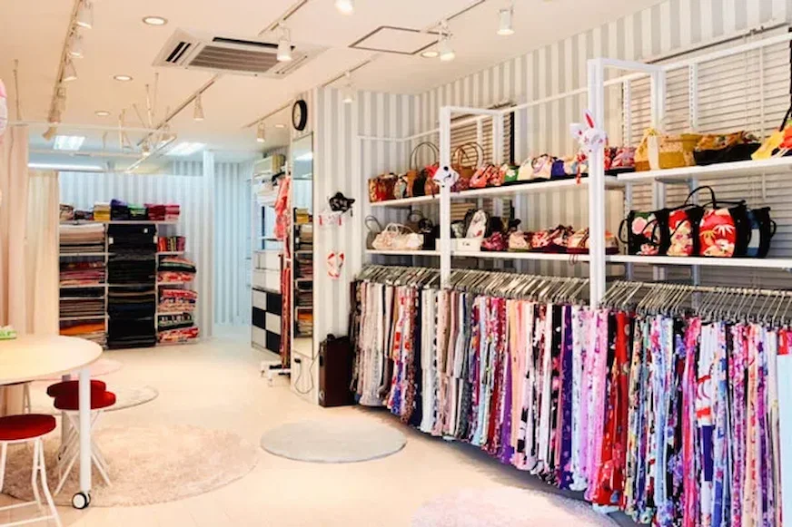 服飾店內景,牆上掛滿色彩豐富的布料和包包,左側有白色桌椅,背景為淺色條紋牆面,環境整齊明亮