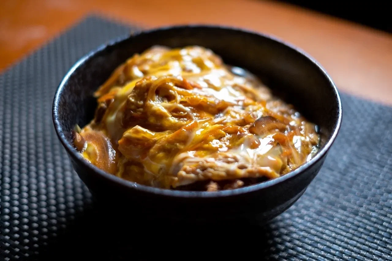黑色碗中盛著金黃色醬汁的蓋澆飯,碗放在深色紋理的桌布上,背景色調溫暖,食物顯得美味可口
