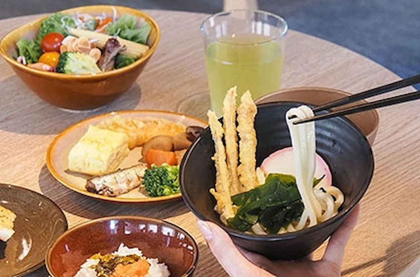 一隻手用筷子夾著碗中烏龍麵,背景有沙拉、飲料和各種日式菜餚,色彩豐富,氛圍輕鬆