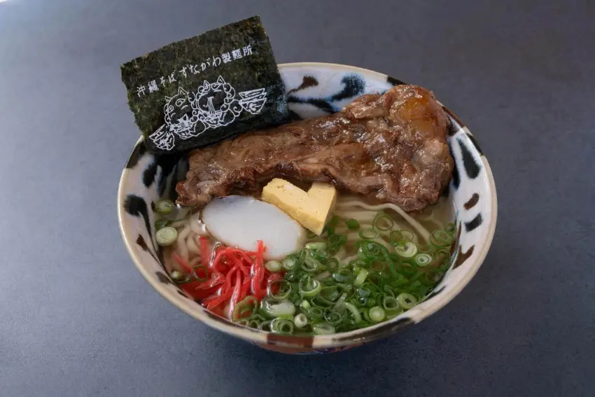 一碗日式拉麵,面上有大塊烤肉、半熟蛋、蔥花、紅薑和海苔,海苔上有日文標識,置於深色桌面,食物看起來豐盛誘人