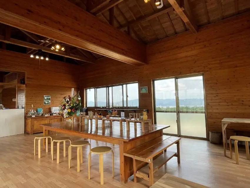 木質大廳內擺著長桌和小凳,桌上有花藝與餐具,落地窗外可見群山與天空,空間明亮寧靜